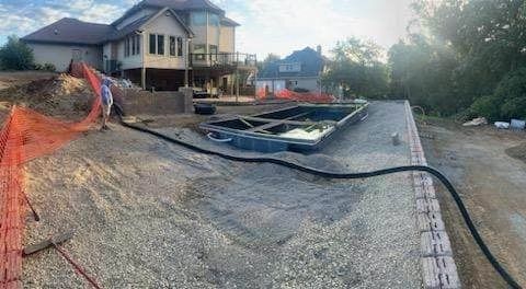 Construction site with a house, exposed driveway, gravel, orange fencing, and a trench line along the curb