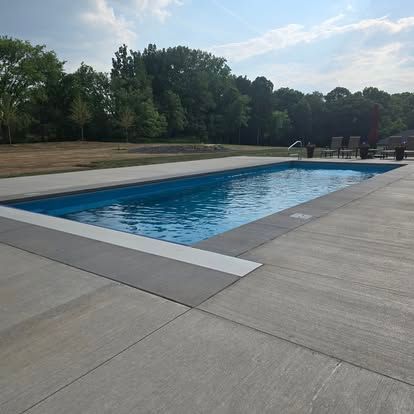 Outdoor rectangular swimming pool beside a concrete deck and trees under a partly cloudy sky