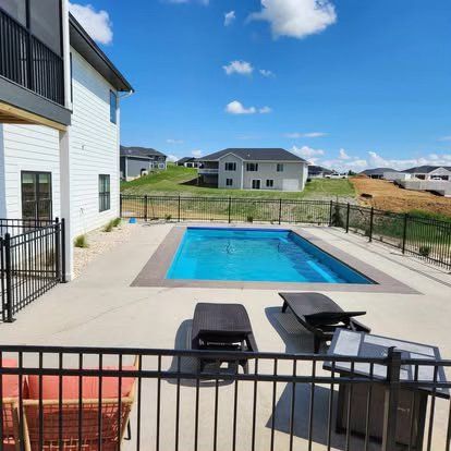 Backyard patio with a small blue swimming pool, black fence, and lounge chairs beside a house.