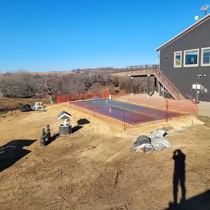 New home construction site with a foundation slab, orange safety fencing, and a house at right.