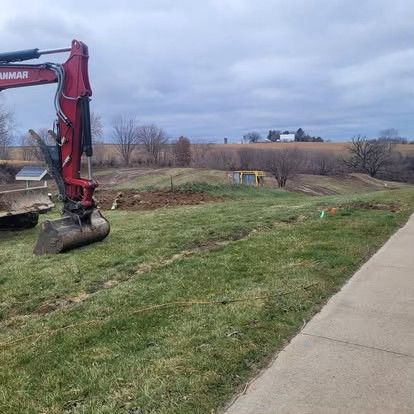 Red excavator beside a grassy slope and sidewalk under a cloudy sky.