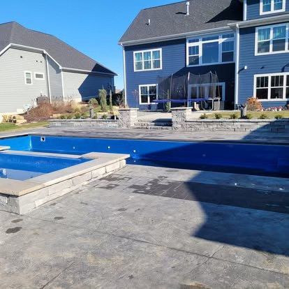 Backyard patio with a blue pool beside a two-story house on a sunny day
