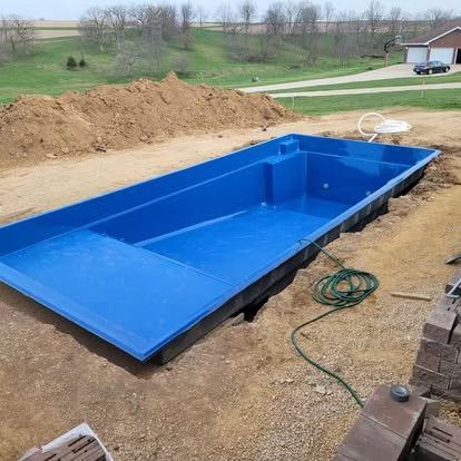 Blue fiberglass pool shell set in a dirt construction site with tools and a mound of soil nearby.