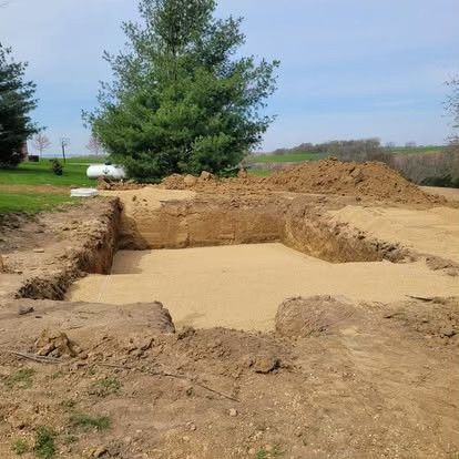 Rectangular excavation filled with concrete, surrounded by dirt piles and a tree in a rural field