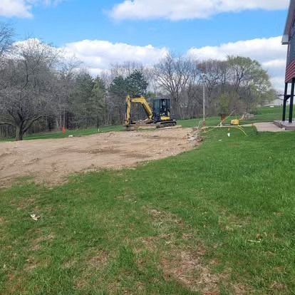 Yellow excavator leveling dirt beside a grassy yard under a partly cloudy sky
