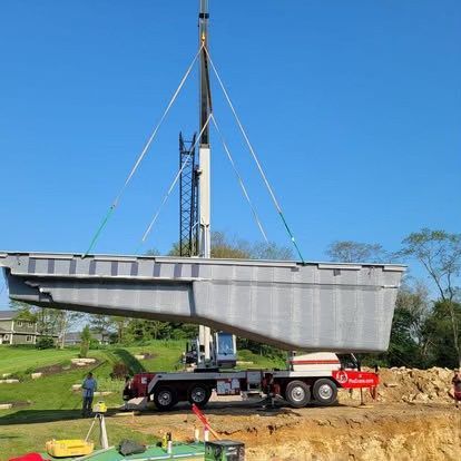 Crane lifting a large gray bridge section over a grassy construction site under a blue sky