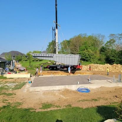 Construction crew lifting a gray dump trailer with a crane over a foundation slab on a sunny lot