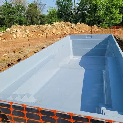 Empty rectangular in-ground pool with light blue lining at a construction site, surrounded by dirt and trees