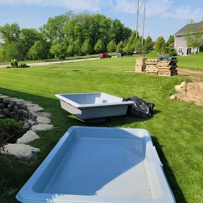 Two white fiberglass tubs on a grassy lawn beside a stone wall, with a house and stacked pallets in the background