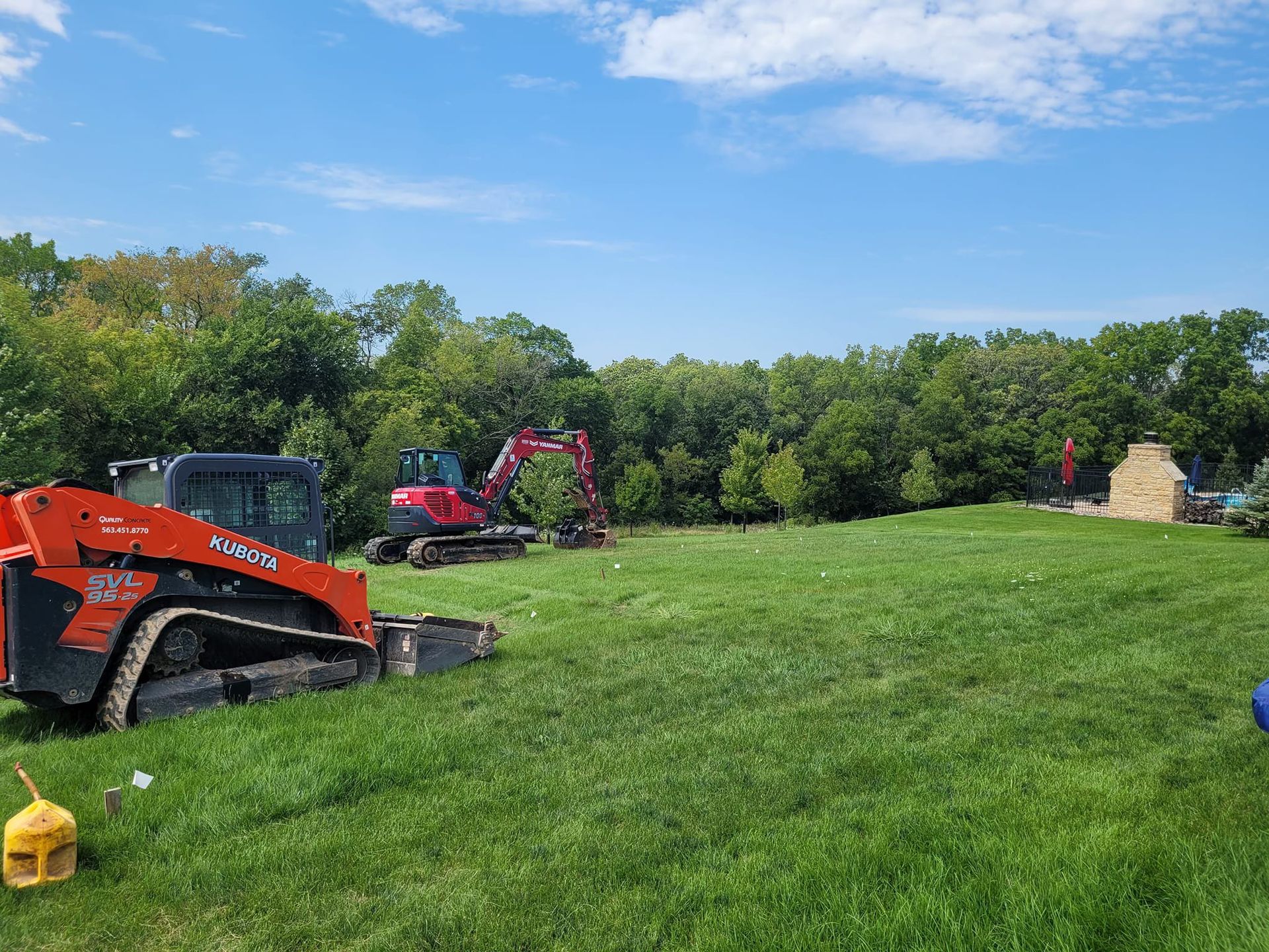 Orange excavator and red mower on a grassy field beside trees under a blue sky.