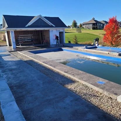 Backyard pool under construction beside a white garage, with gravel borders and one worker nearby