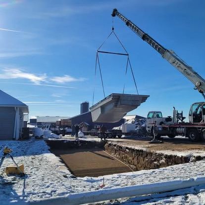 Crane lifting a large metal structure over a snowy construction site beside a trench