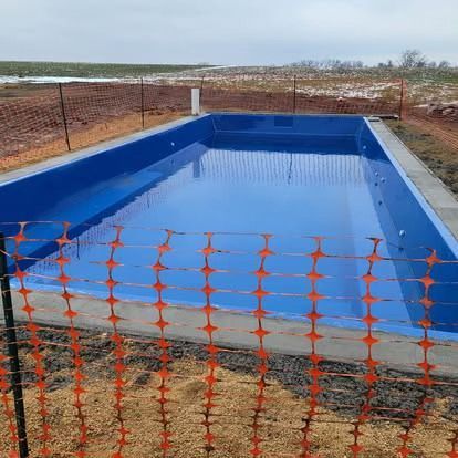 Blue-lined outdoor swimming pool under construction, enclosed by orange safety fencing on a dry dirt site.