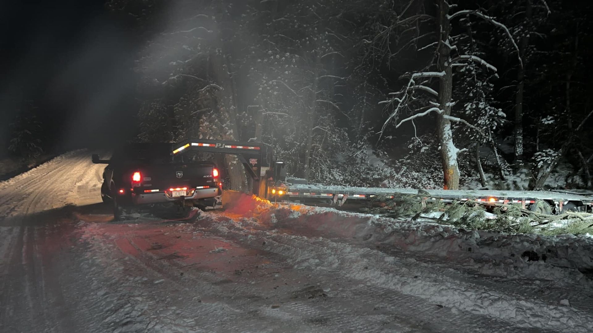 Truck stuck on a snowy road at night, with two people and smoke. Trees and guardrail are visible.