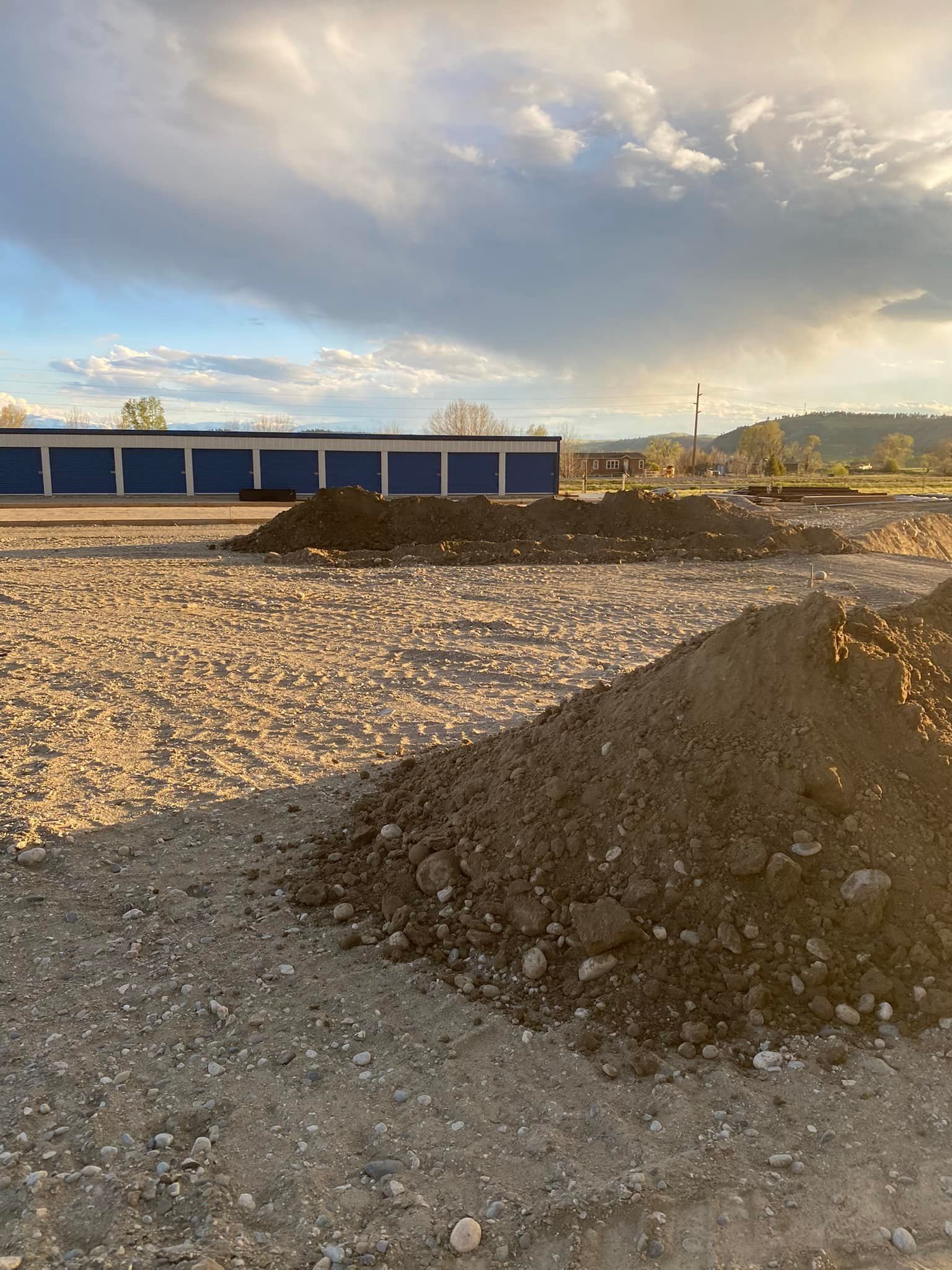 Dirt piles in a gravel lot with a blue-walled building in the background under a cloudy sky.