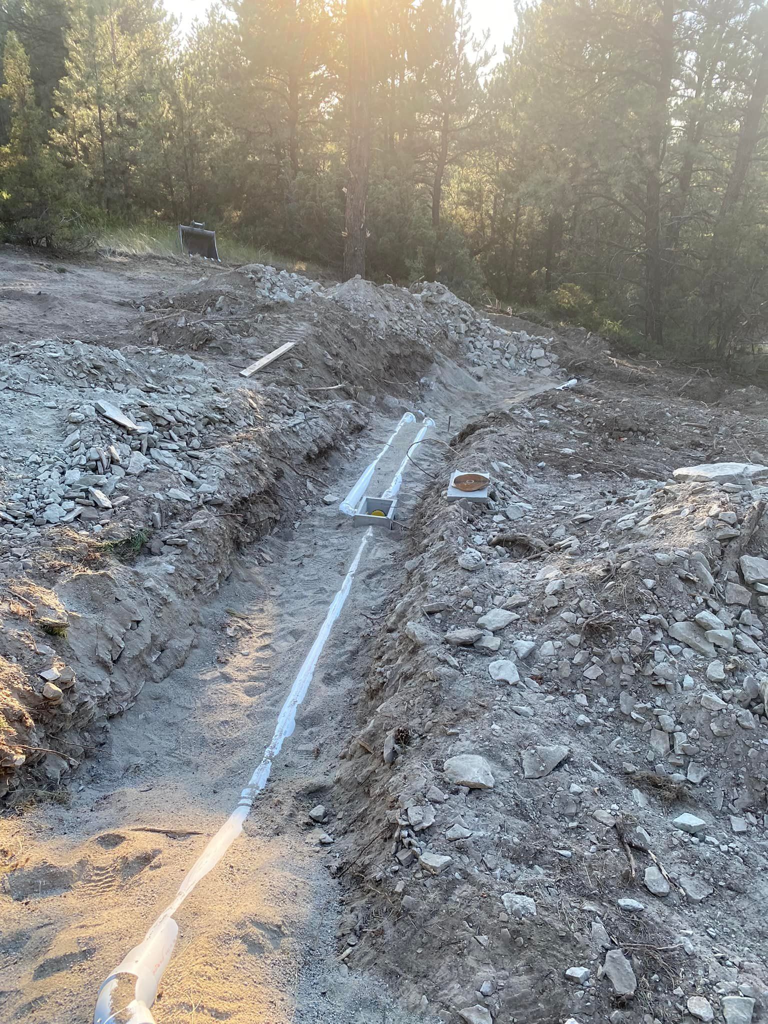 Trenches with white pipes laid, in a dirt and rock construction site with trees and sunlight in the background.