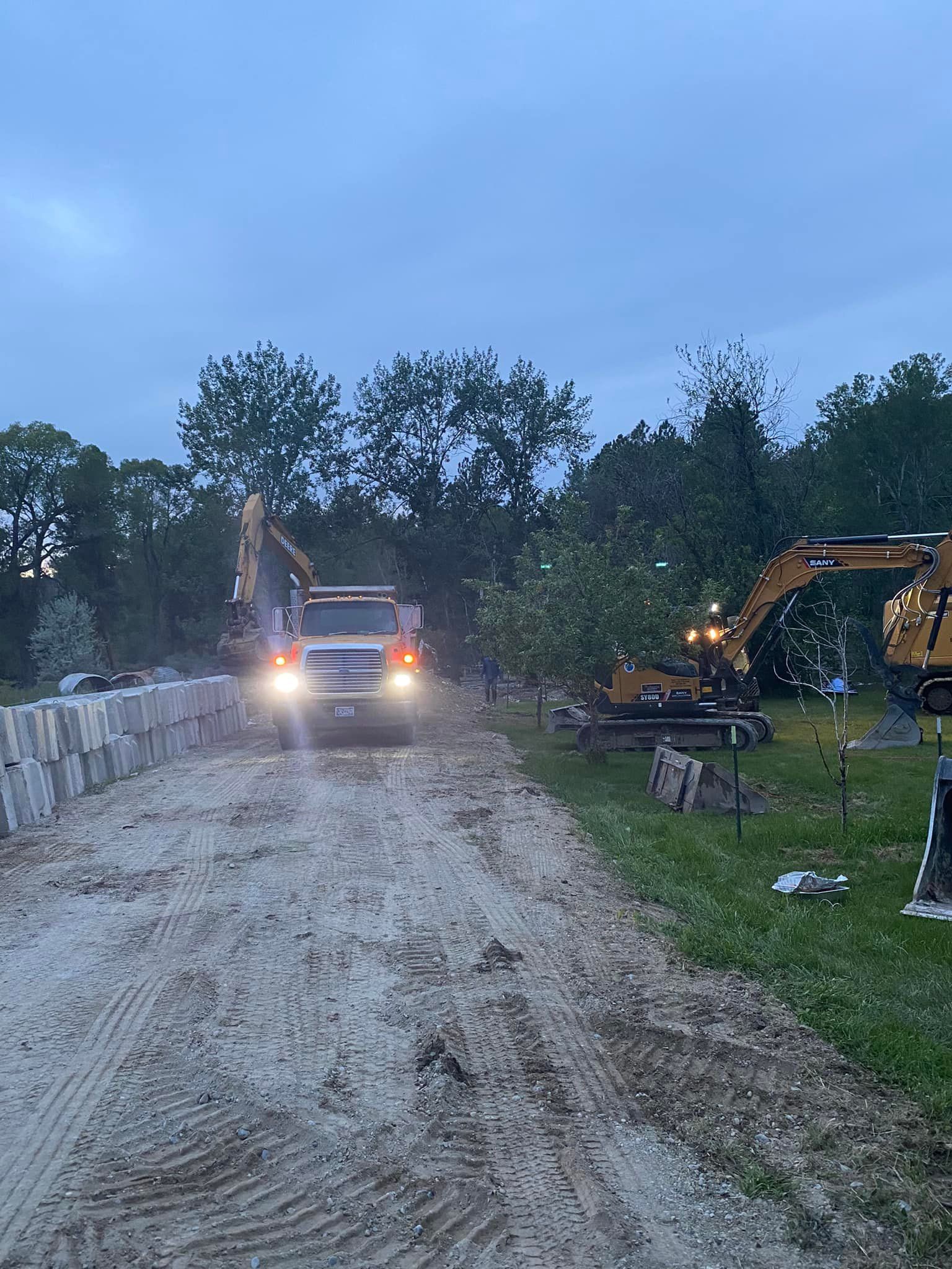 A truck drives on a dirt path with two excavators nearby. Gray retaining wall on the left, trees in the background.