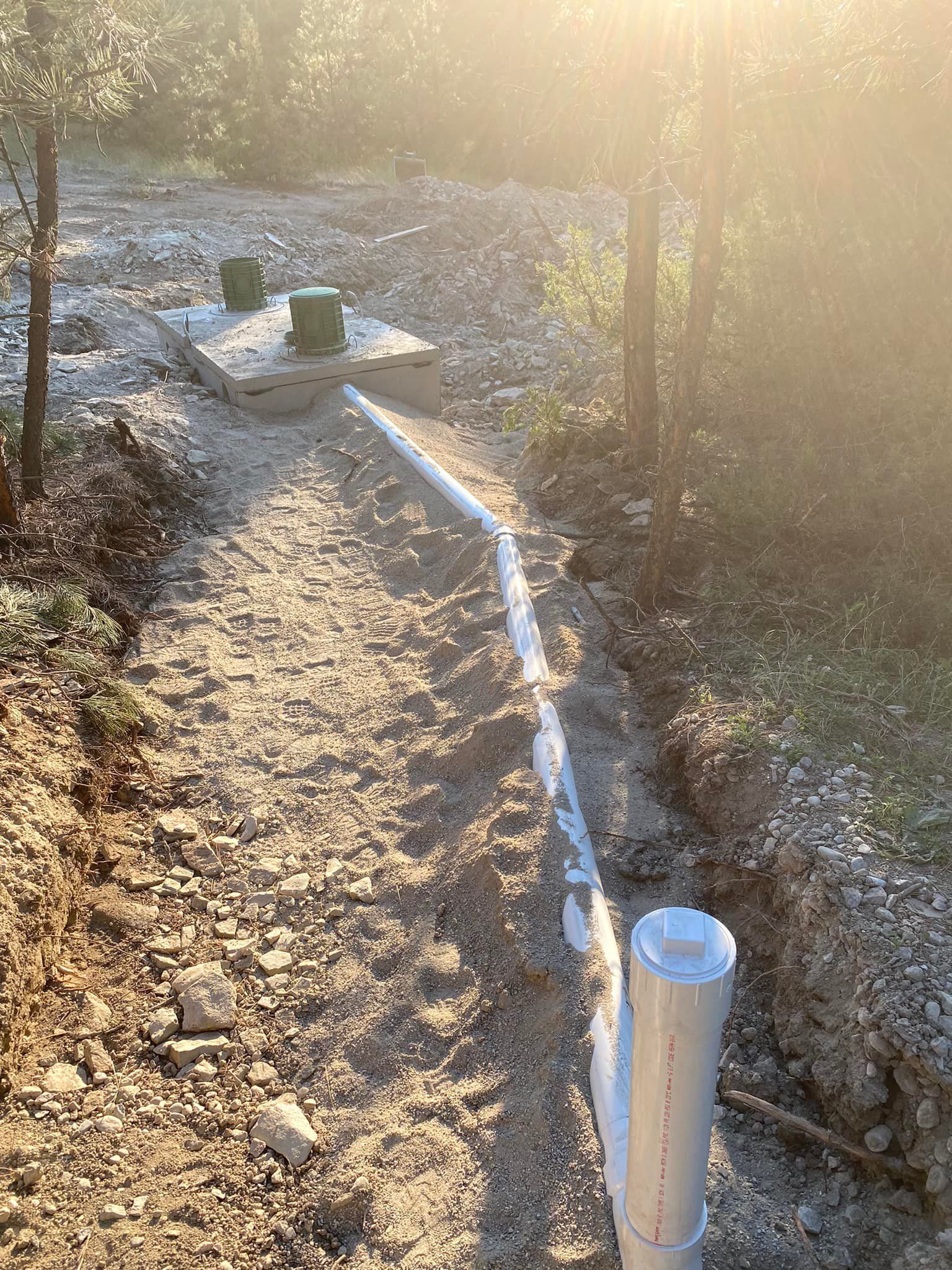 Gravel pathway with white pipes and a concrete septic tank in a wooded area, lit by sunlight.