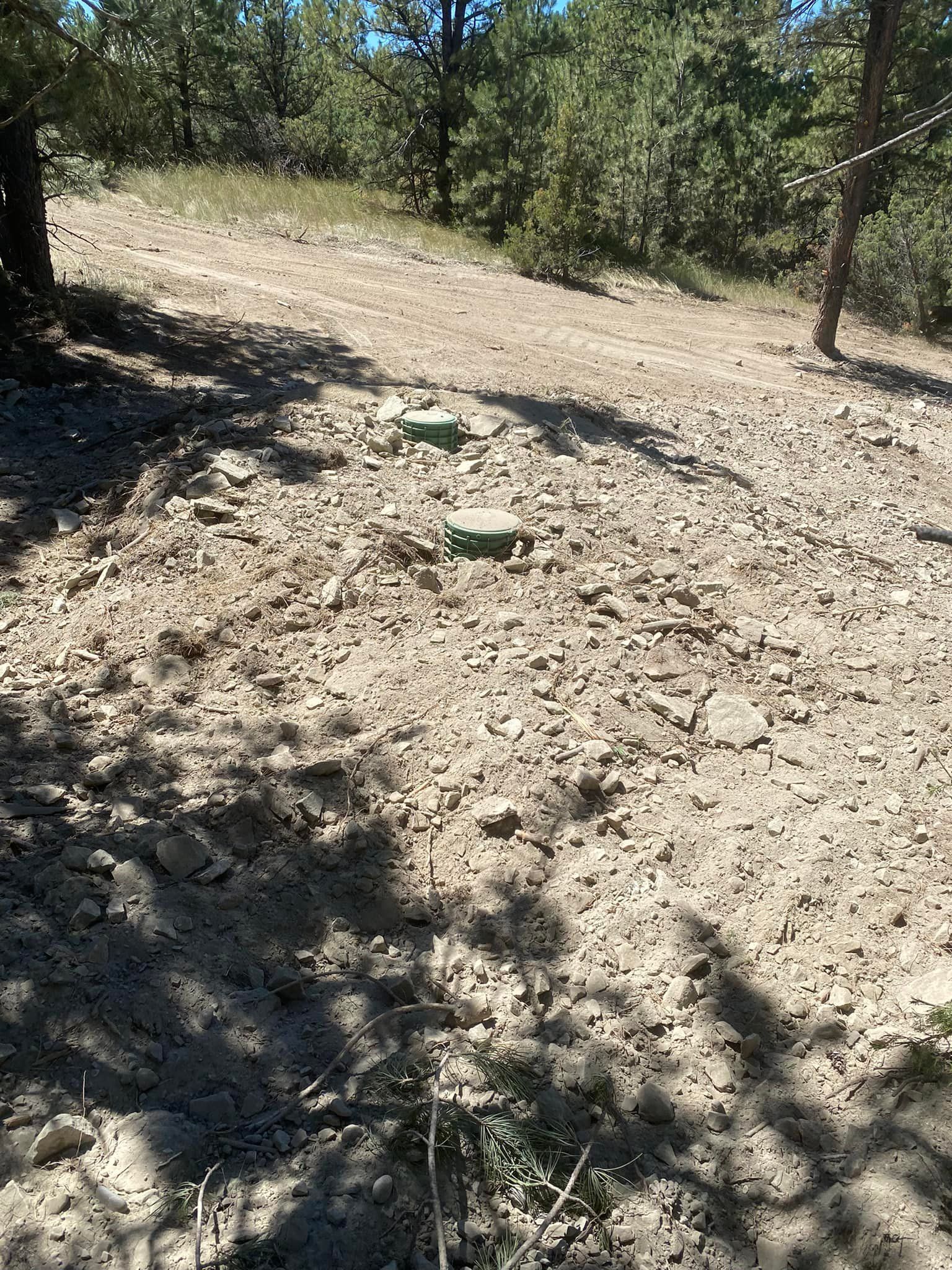 Three green capped pipes in dry, sandy ground, on a hillside with trees and bright sunlight.