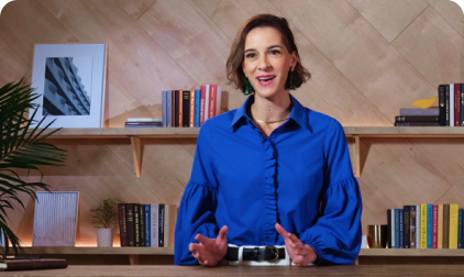 Woman in blue shirt speaking, gesturing with hands; shelves with books and art in the background.