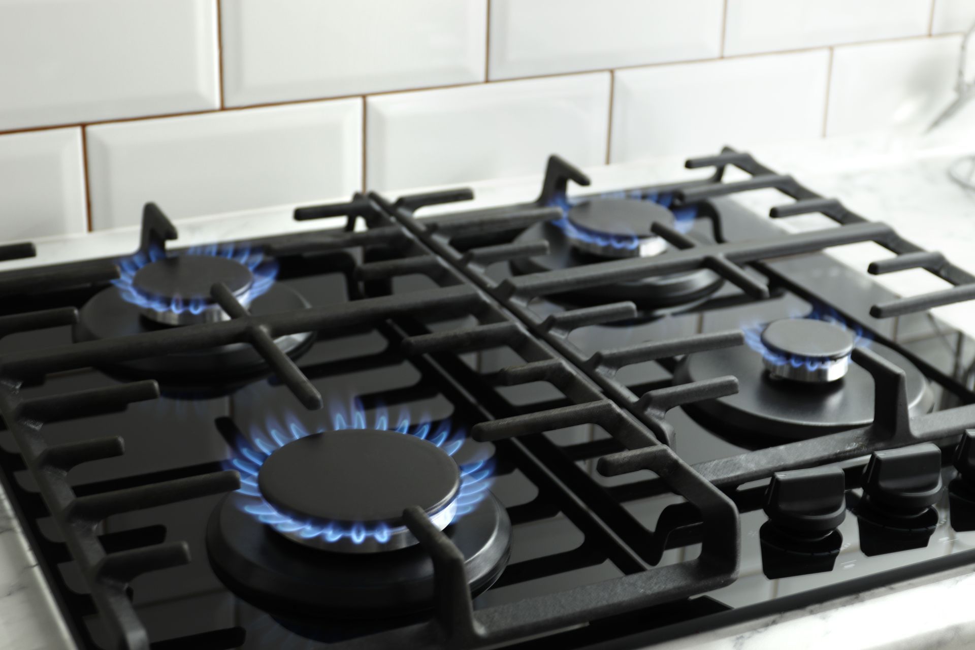 Gas cooktop with blue flame burners lit on a black stovetop against white tile