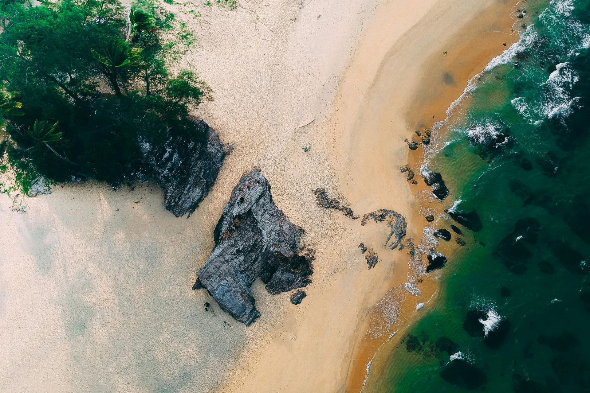 Aerial view of a sandy beach with dark rocks beside turquoise ocean water and green vegetation