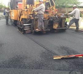 Road paving machine laying fresh asphalt while workers stand beside it on a wet street