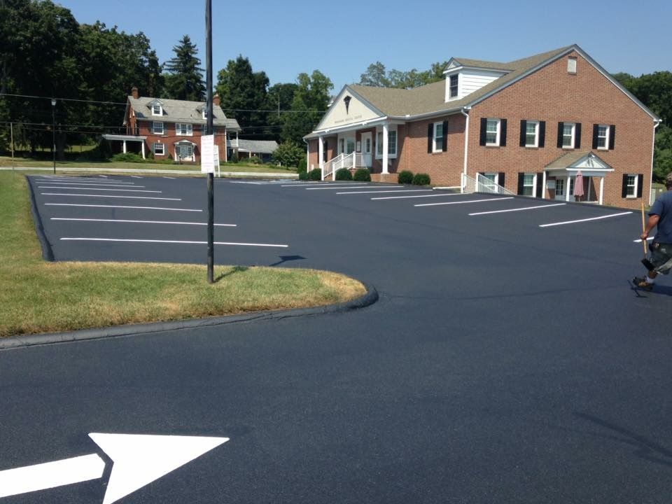 Freshly paved parking lot beside brick buildings, with white directional arrows and a grassy island.