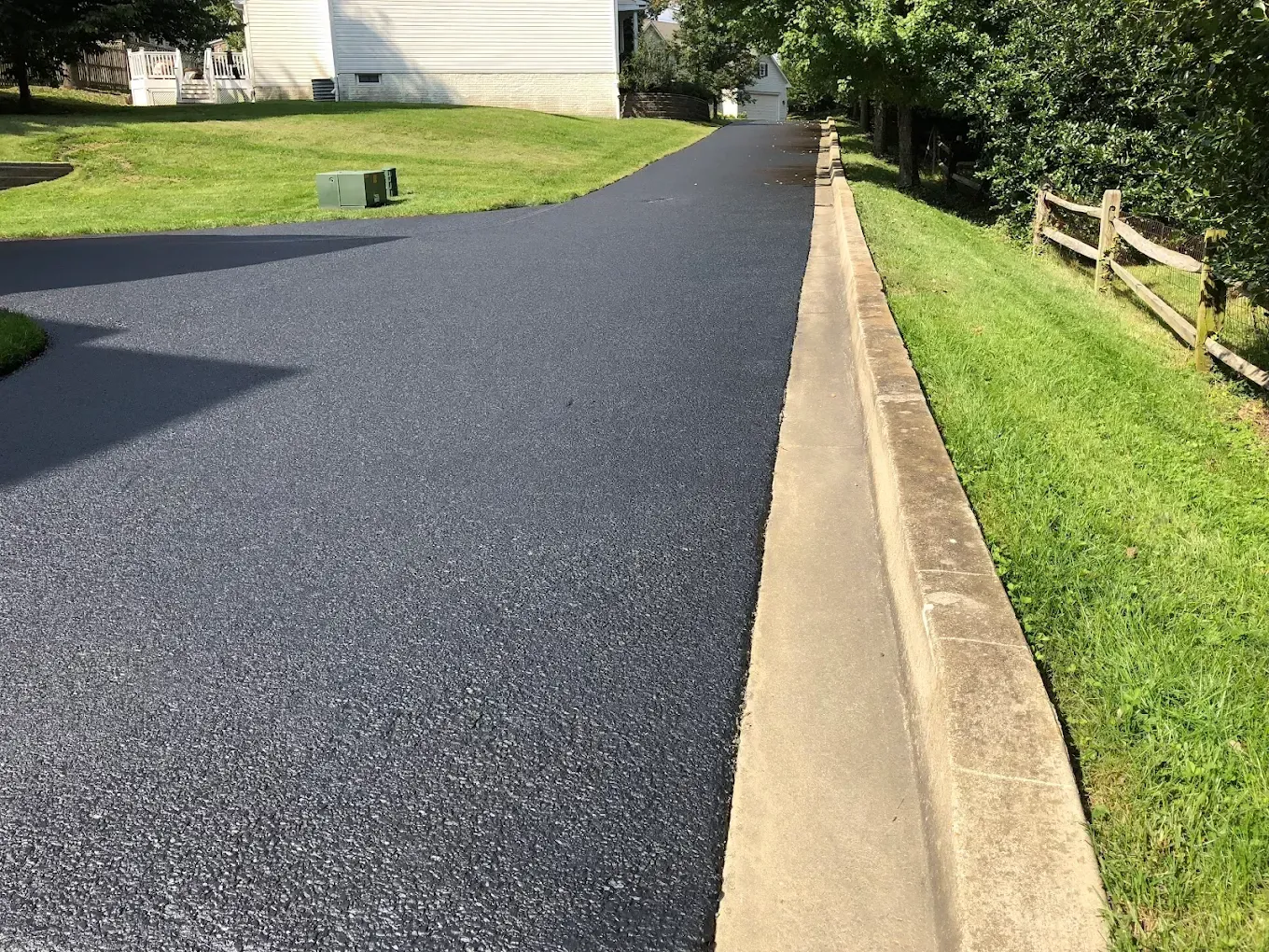 Freshly paved black driveway beside a concrete curb, with grass and trees along the edge