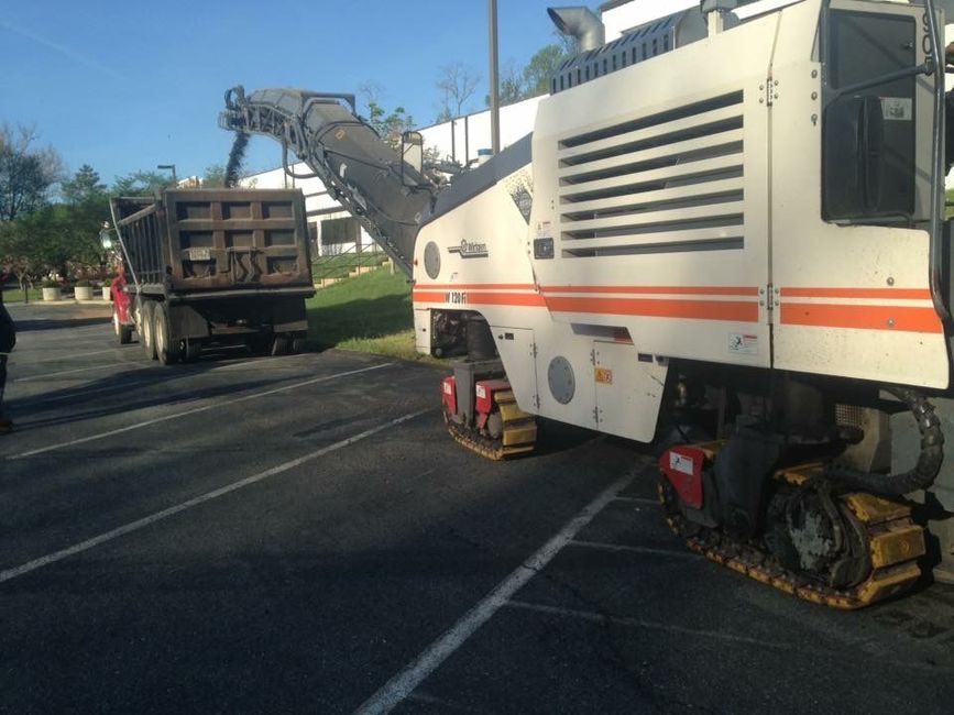 White road milling machine beside a brown dump truck on a sunny street, with a worker nearby
