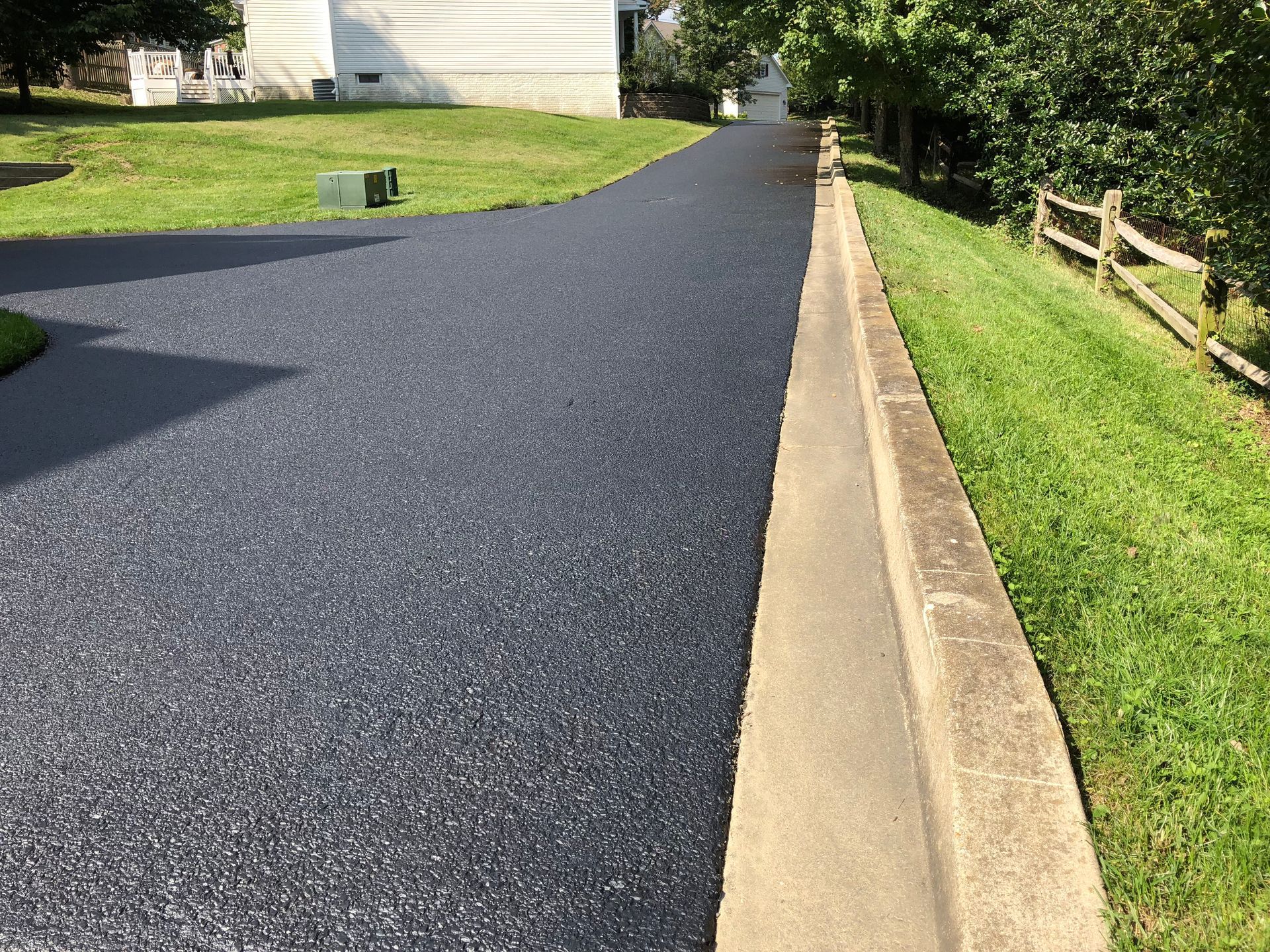 Freshly paved black driveway beside a tan curb and green lawn, leading toward a house and trees