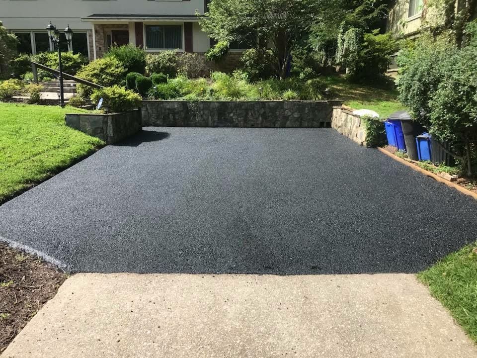 Freshly resurfaced black asphalt driveway bordered by stone walls and greenery in a suburban yard