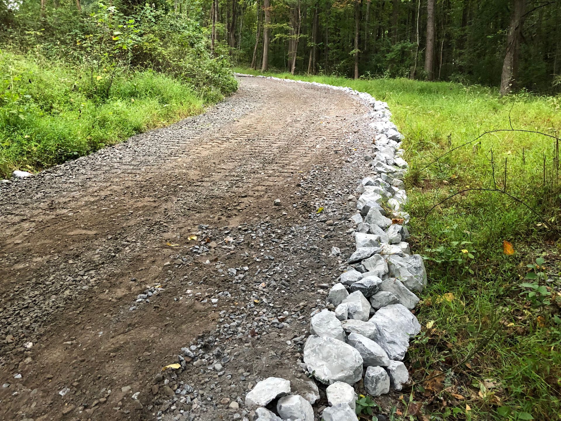 Curving gravel road through a green forest, lined with large rocks on one side.
