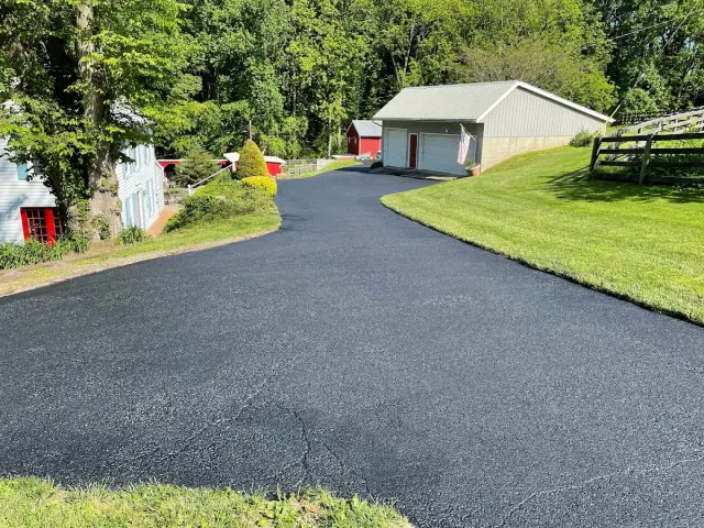 Freshly paved black driveway curving toward a gray garage beside a grassy yard and trees