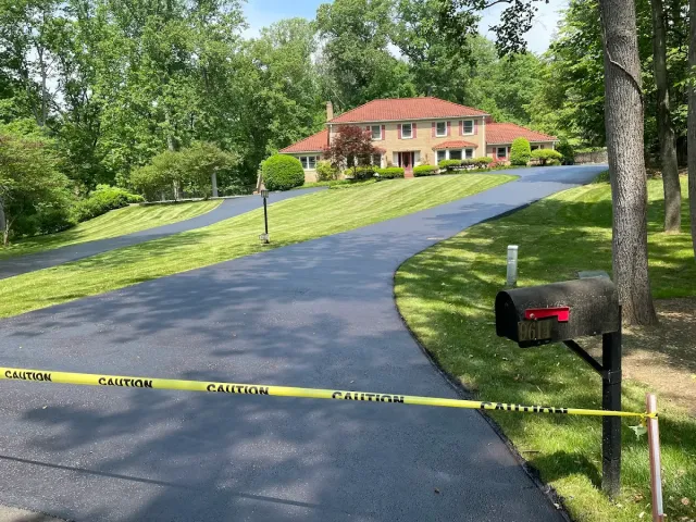 Police tape blocks a long driveway to a large house surrounded by trees and lawn.