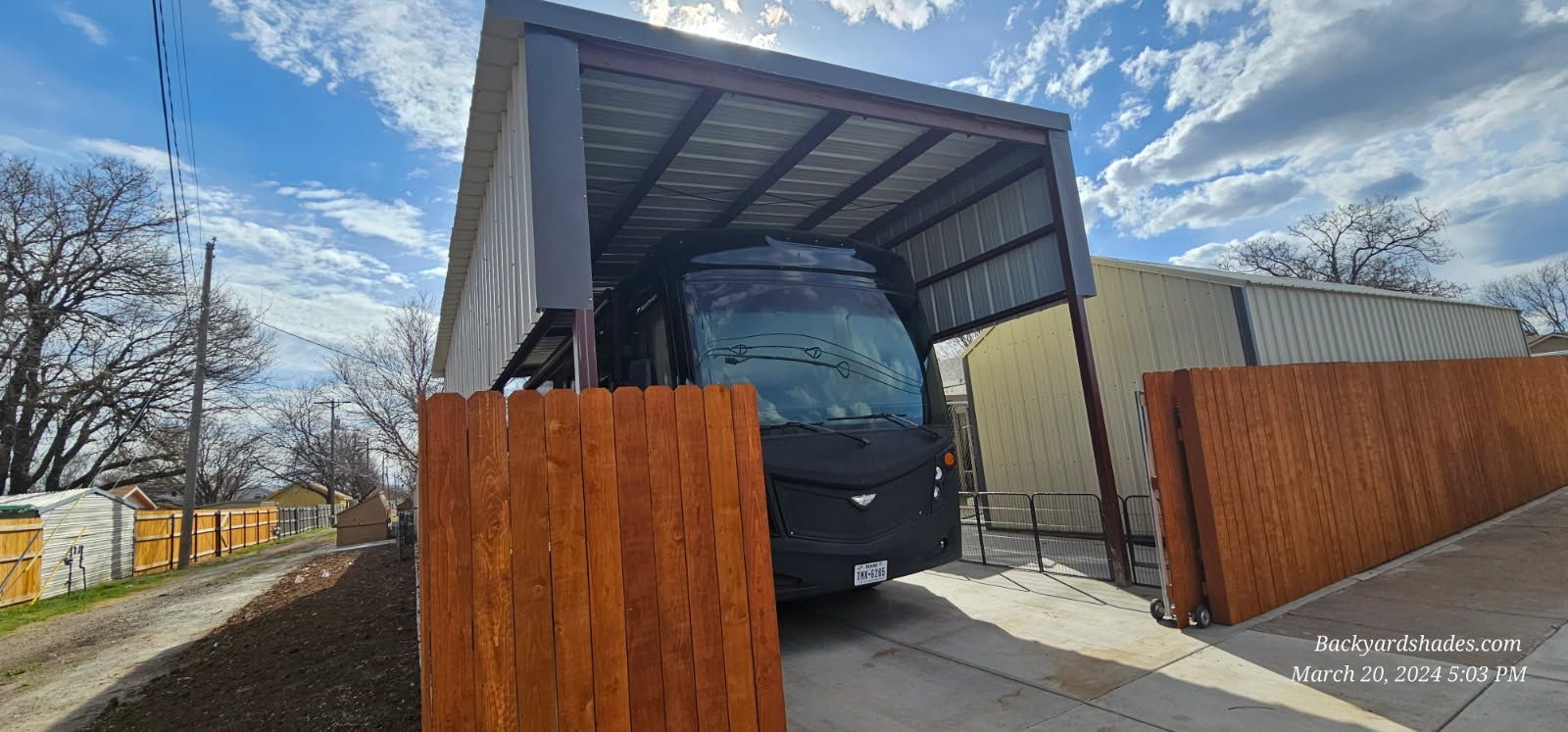 A large metal carport with a brown roof and supports, standing next to a garage, under a blue sky.