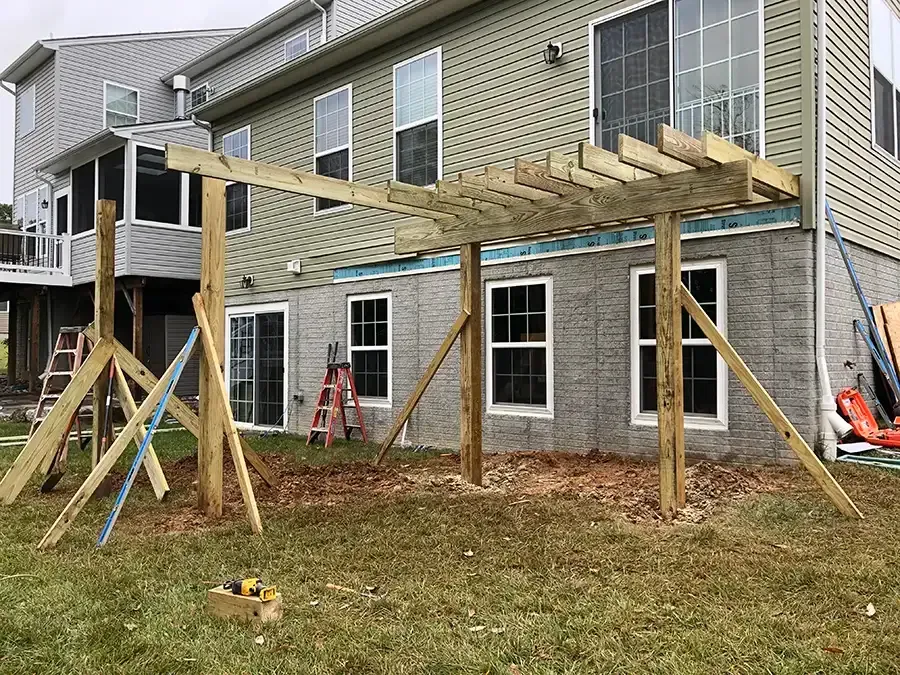 Wooden pergola frame under construction beside a townhouse, with support braces and ladders in a yard.