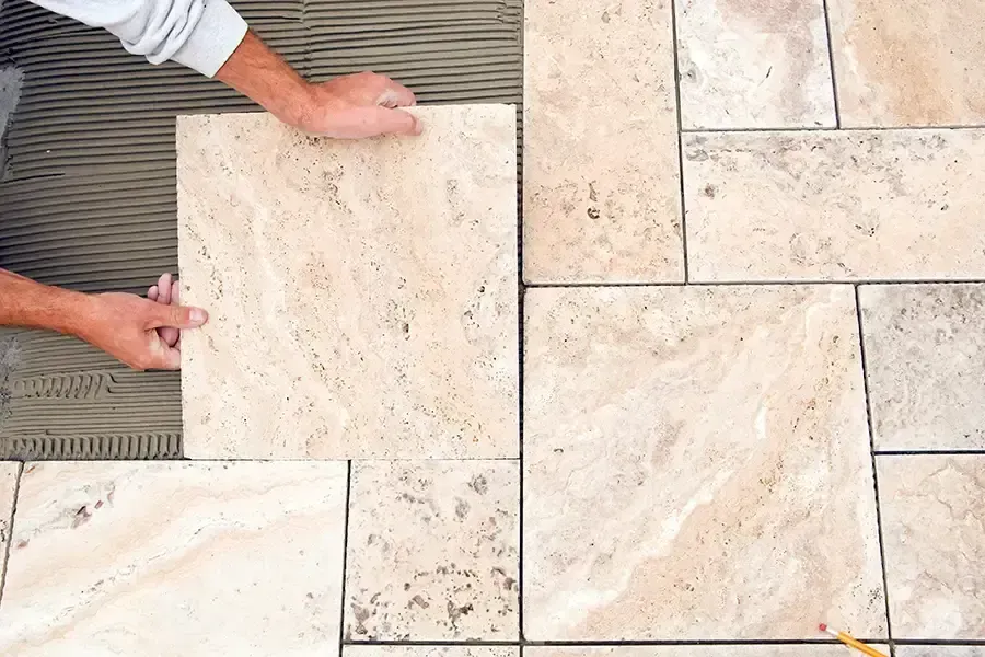Hands laying a beige stone tile on a patio beside already installed tiles.