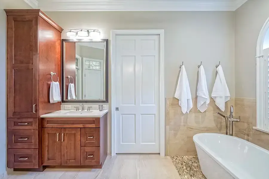 Bright bathroom with wooden vanity, white door, towels, and a bathtub with beige tile.