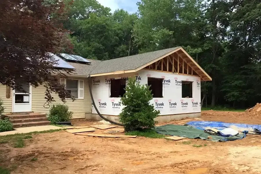 Partially built house with white siding, exposed roof framing, and construction materials in a wooded yard