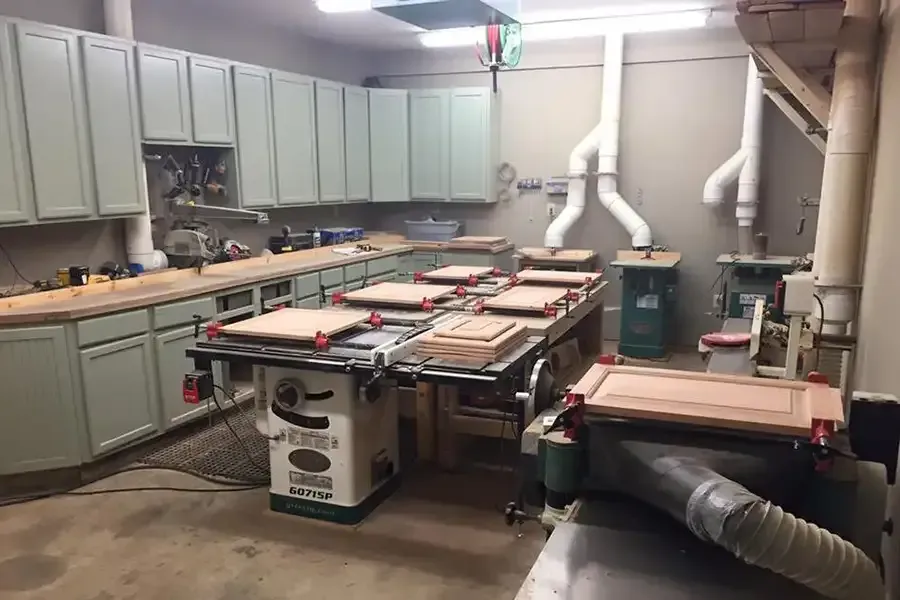 Workshop with saw table, cabinets, and dust-collection pipes in a basement woodworking shop.