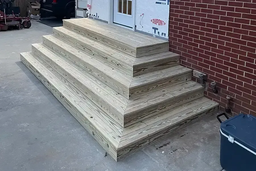 Wooden stepped platform beside a brick building entrance, with concrete ground and a trash bin nearby
