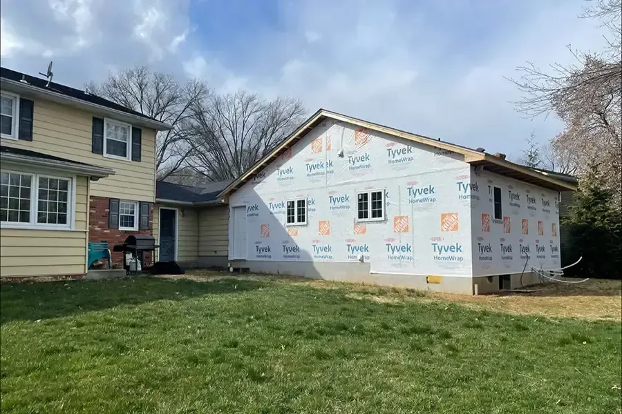 House under construction with white house wrap beside an existing beige home and grassy yard