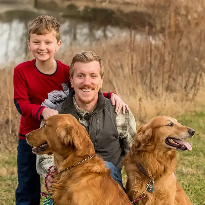 Smiling man and boy outdoors with two golden retrievers in a grassy field