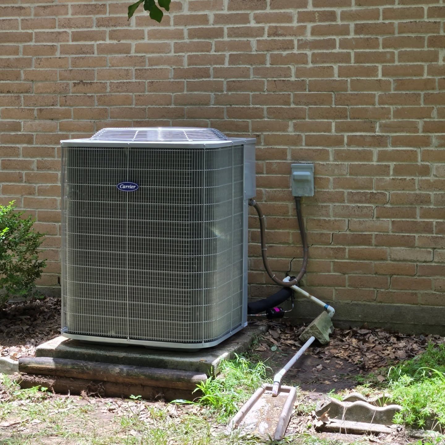 Outdoor air conditioner unit beside a brick house wall with garden hose and electrical conduit nearby