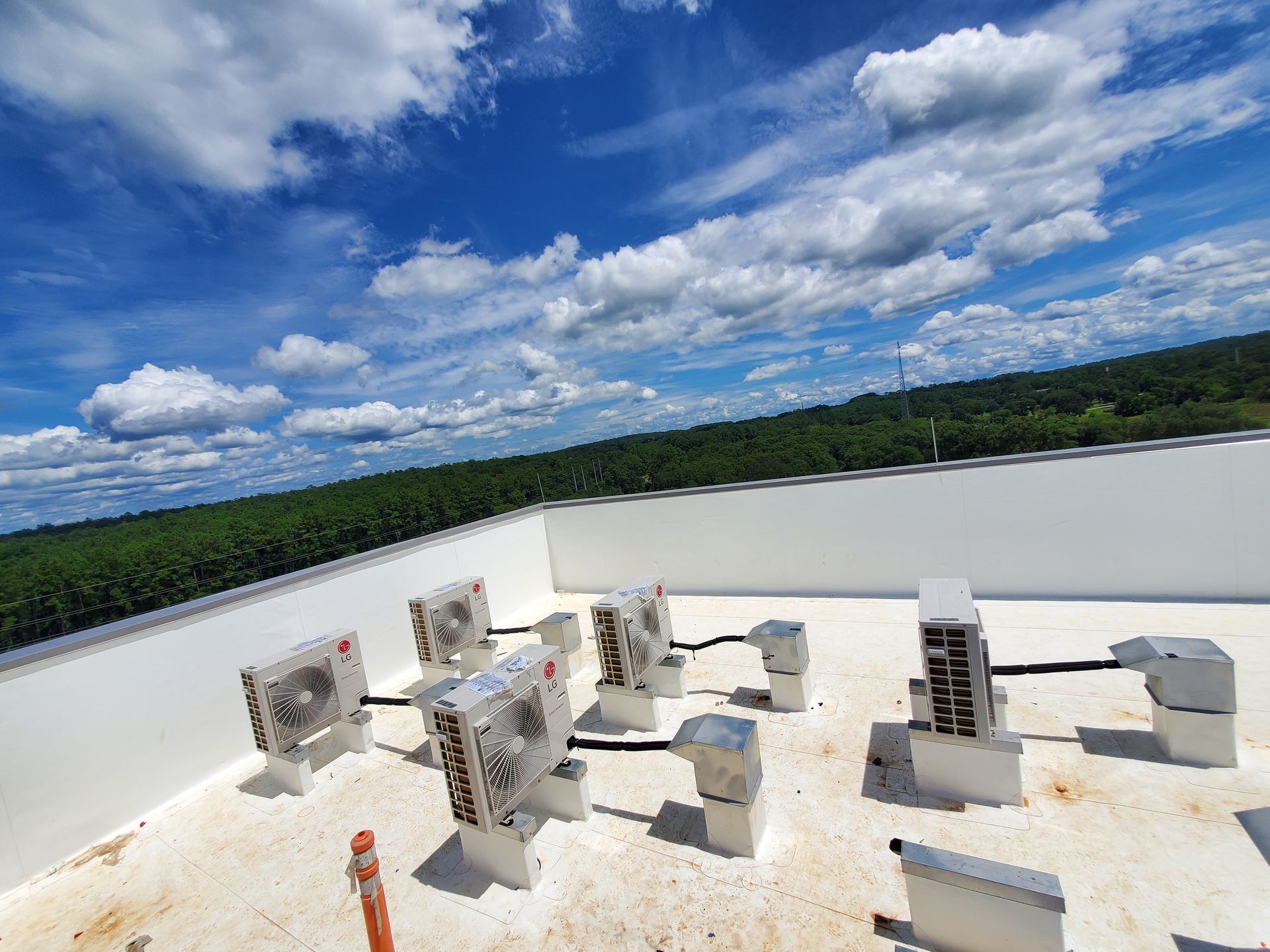 Rooftop with several air conditioning units against a backdrop of trees and a bright, cloudy sky.
