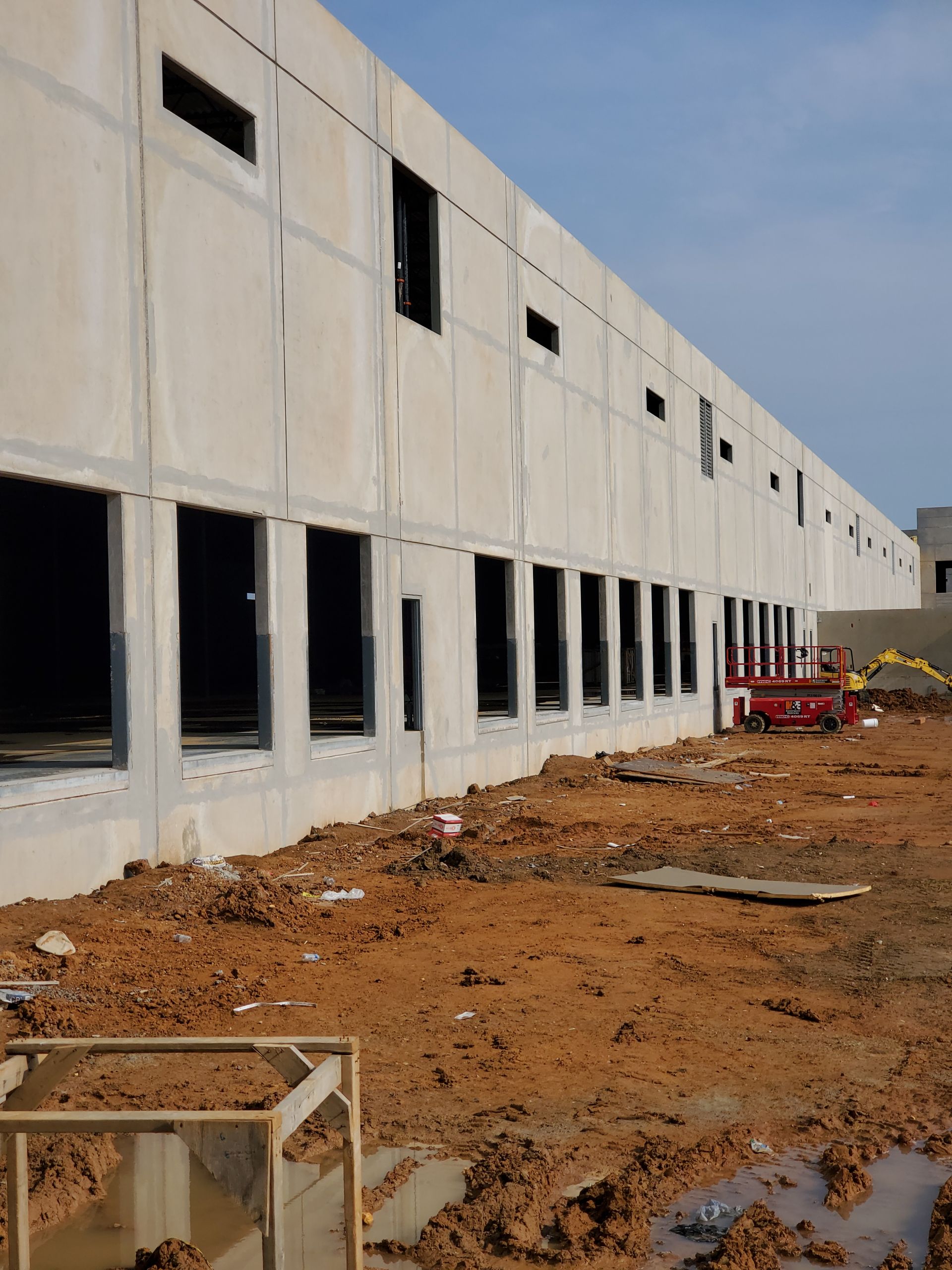 Exterior of a building under construction with concrete walls and empty window frames; muddy ground in the foreground.