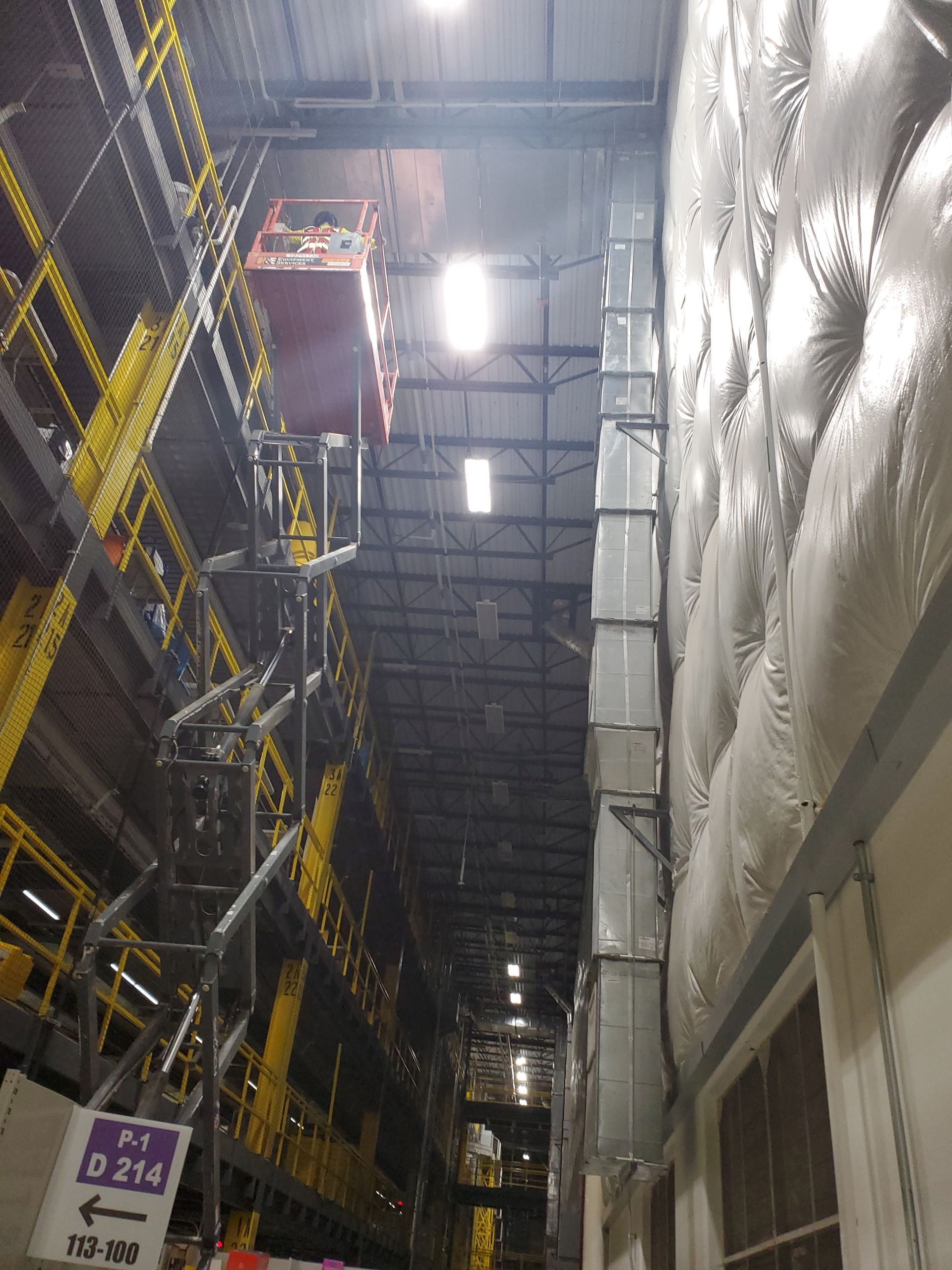 Warehouse interior: A person in a lift near shelves. Silver ductwork runs along a padded wall.