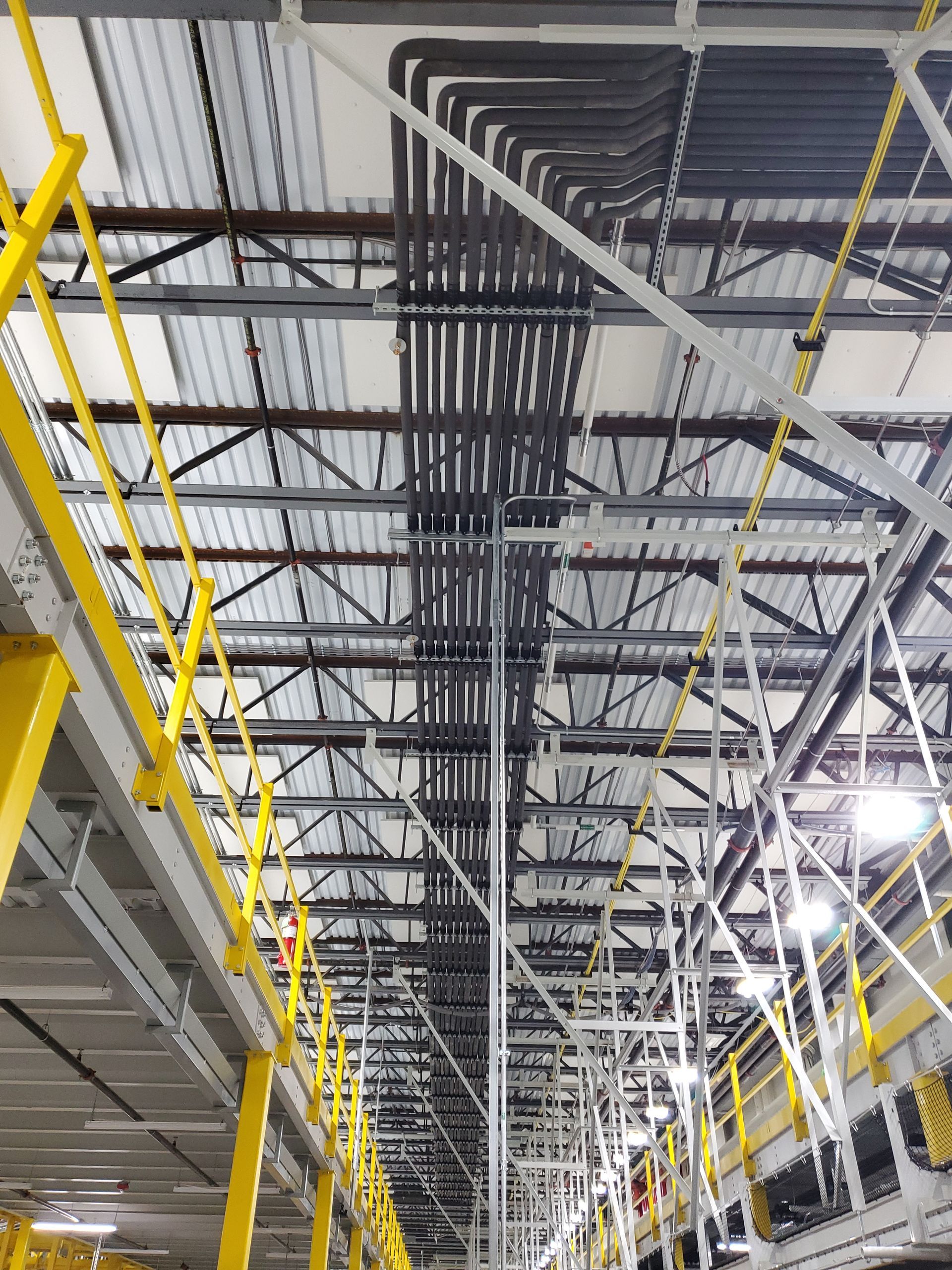 Overhead view of a factory ceiling, showing black electrical conduits, metal beams, and yellow safety railings.