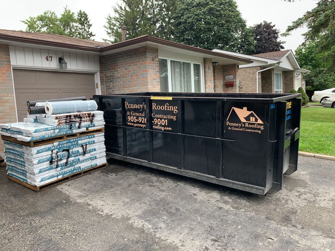 Black dumpster and roofing materials in front of a house, likely for a roof replacement project.